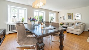 A spacious living room with a large wooden dining table, wicker chairs, a beige sofa, and a chandelier.