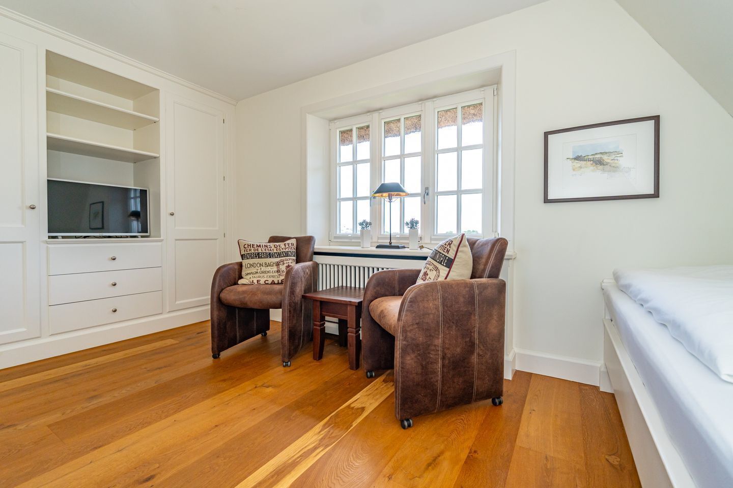 Une pièce avec un plancher en bois, un bureau blanc avec une lampe, deux fauteuils marron, un lit blanc et un tableau encadré au mur.