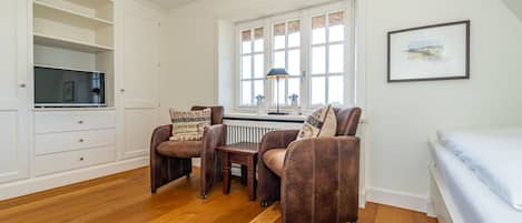 A room with a wooden floor, a white desk with a lamp, two brown armchairs, a white bed, and a framed picture on the wall.