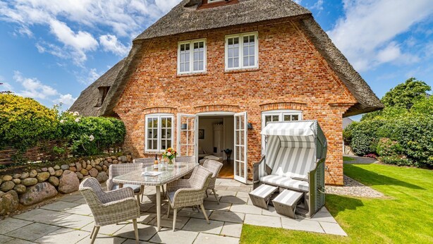 A brick house with a thatched roof, a patio with wicker furniture, and a well-maintained garden.