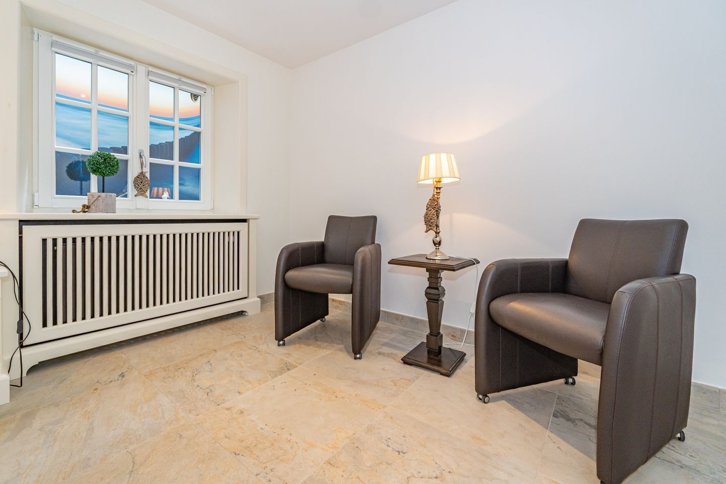 Een kamer met twee leren fauteuils, een tafeltje met een lamp, een radiator en een raam met uitzicht op zee.