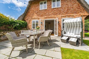 A brick house with a thatched roof, a glass table, wicker chairs, and a grill outside.
