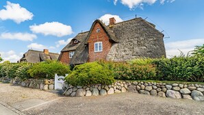 A traditional stone house with a thatched roof, surrounded by a stone wall and lush greenery. - Hüs bi Koog 1 - Vacation under thatch for up to 5 people. (Sylt)
