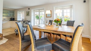 A dining area with a wooden table and black leather chairs, a kitchen in the background, and a window with a view of greenery. - Hüs bi Koog 1 - Vacation under thatch for up to 5 people. (Sylt)