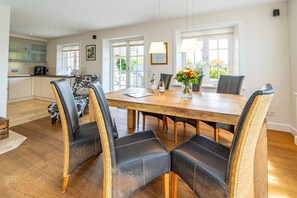 A dining area with a wooden table and black leather chairs, a kitchen in the background, and a window with a view of greenery. - Hüs bi Koog 1 - Vacation under thatch for up to 5 people. (Sylt)