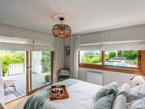 Ett sovrum med en säng, ett brickbord med frukostmat, en stol och utsikt över en swimmingpool och grönområden utanför.