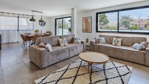A modern living room with two sofas, a dining area, and large windows offering a view of the neighborhood. - Denneweiller by Interhome (Denneville)