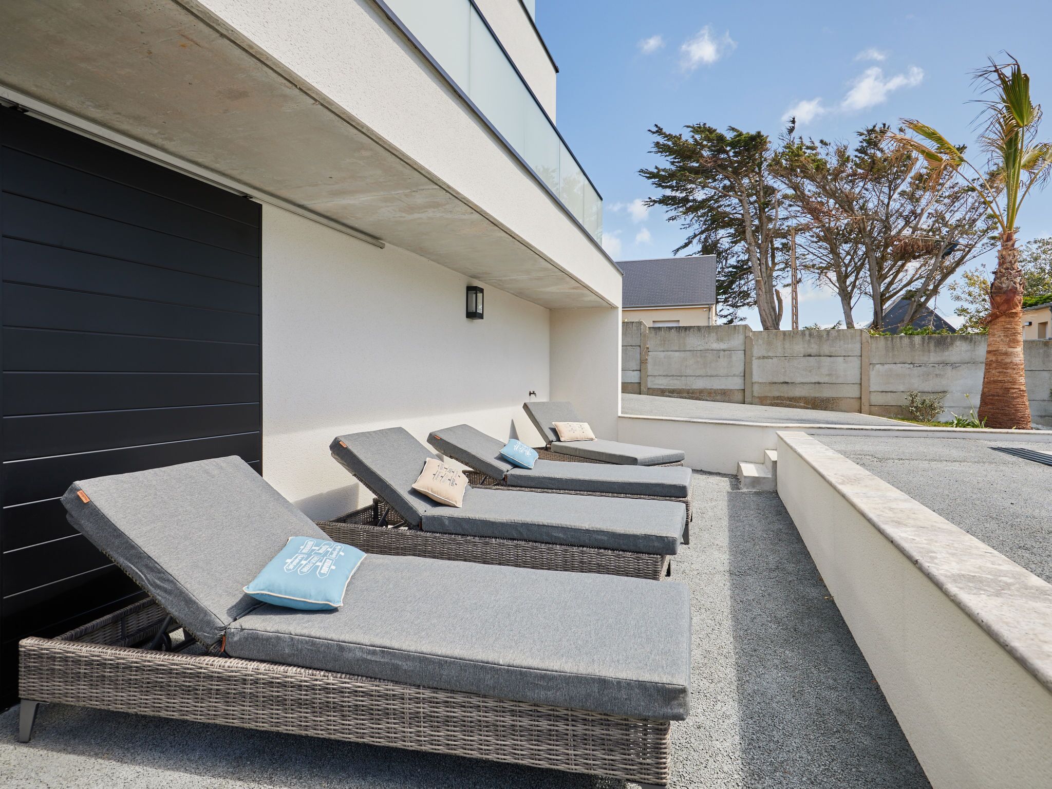 Een moderne buitenlounge met drie loungestoelen, een betonnen muur en een palmboom.