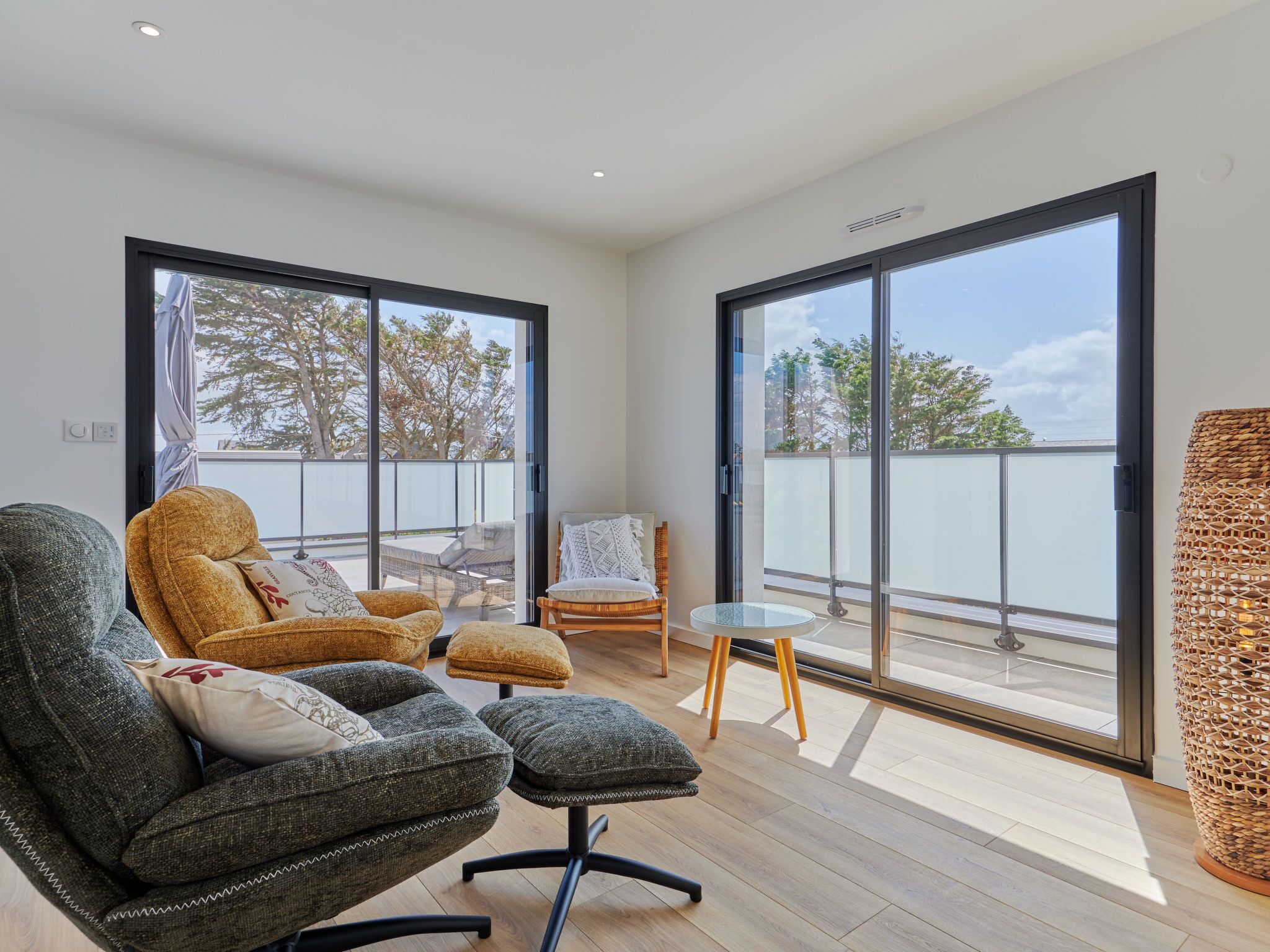 Een moderne woonkamer met een grijze bank, een kleinere fauteuil en een kleine ronde tafel. Grote schuifdeuren leiden naar een balkon met uitzicht op bomen en een hek.