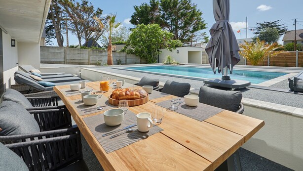 An outdoor dining area with a pool, lounge chairs, and a wooden table set for a meal. - Denneweiller by Interhome (Denneville)