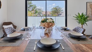 A dining area with a wooden table set for four, a vase of flowers, and a large window with a view of trees and buildings. - Denneweiller by Interhome (Denneville)