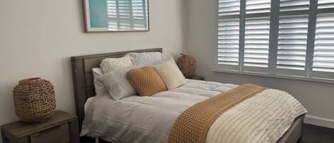 Una cama bien hecha con edredón blanco y varias almohadas, una mesita de noche de madera con una cesta tejida y una ventana con persianas.