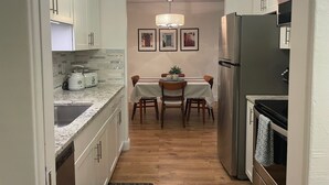 Una cuina estreta amb armaris blancs, un refrigerador d'acer inoxidable, una taula de dinar amb cadires i un lampadari.