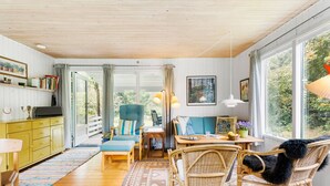 Een gezellige kamer met een houten plafond, een eettafel met stoelen, een rieten fauteuil met een zwart, zacht kussen, en een groot raam met uitzicht op groen.