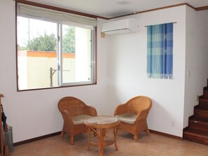 A room with two wicker chairs, a small round table, a window with a view, and a blue patterned wall hanging.
