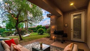 An outdoor patio with a sofa, a coffee table, and string lights. There is a door with a window and a grill in the background. - Lake View 3BR | Resort Pools at Indian Palms (Indio)