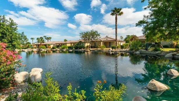 A residential area with a clear blue sky, a small pond, and houses with red-tiled roofs. - Lake View 3BR | Resort Pools at Indian Palms (Indio)
