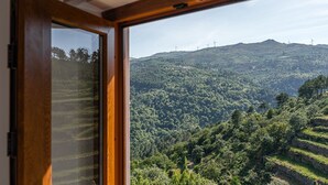 Uitzicht door een raam met houten kader op een weelderig groen landschap met terrassen en windturbines in de verte.