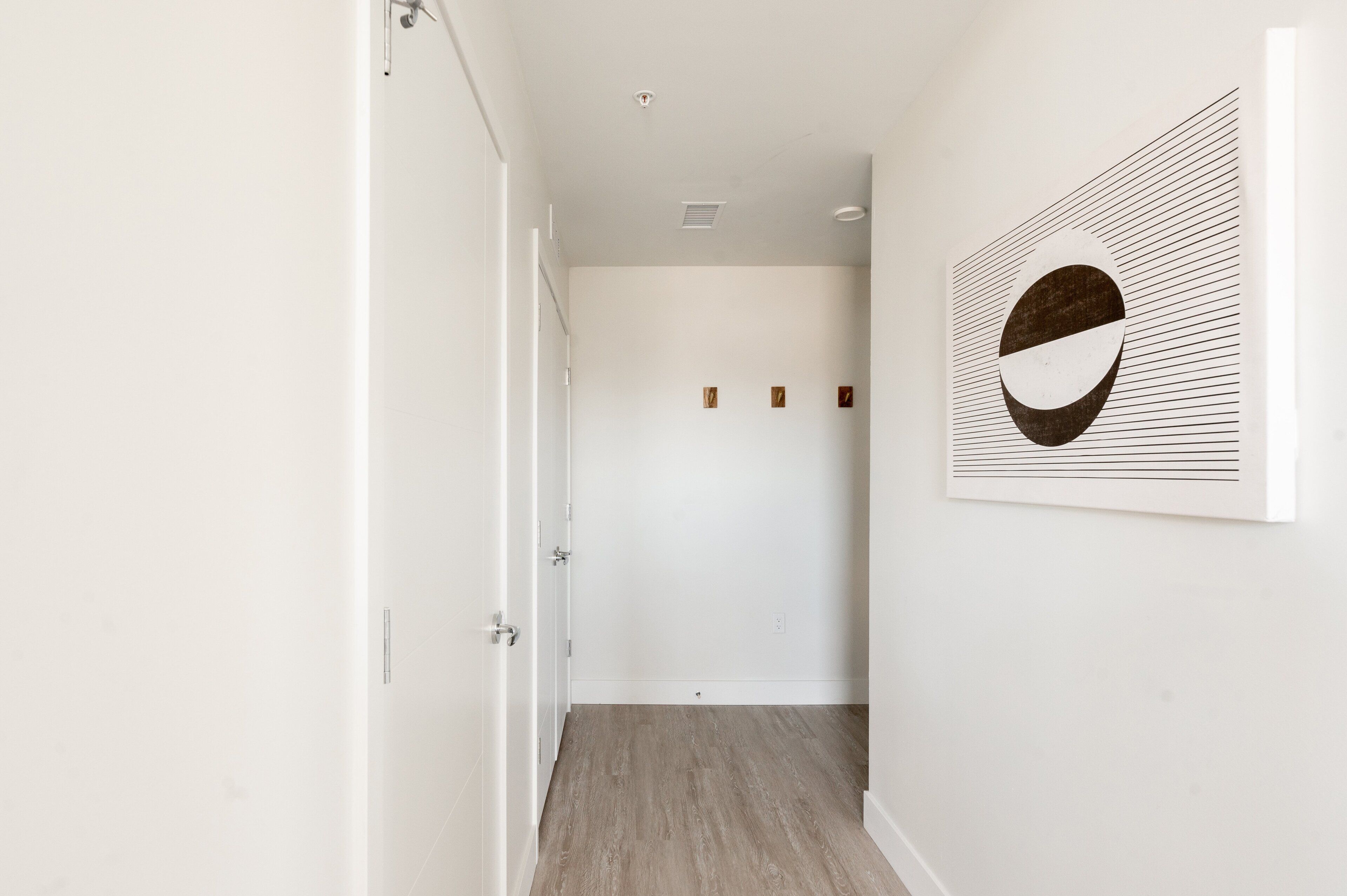 Un pasillo con piso de madera, paredes blancas y una puerta a la izquierda. Se observa una obra de arte colgada en la pared, con un diseño circular y líneas horizontales.