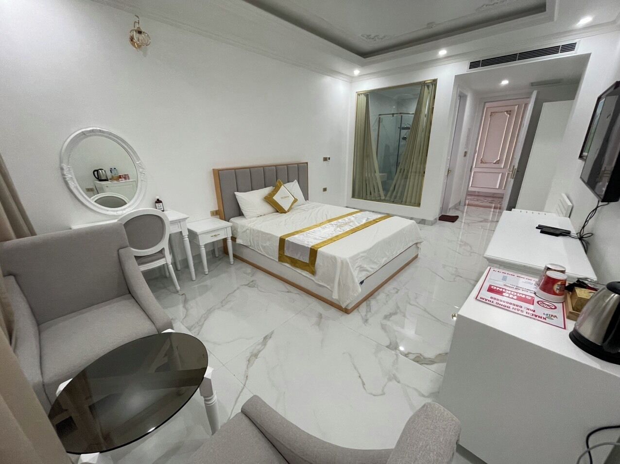 A modern hotel room with a bed, a desk, a chair, a round mirror, and a large window.