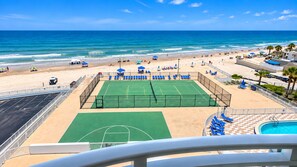 Un court de tennis situé à côté d’une plage, avec une piscine et des parasols.