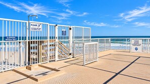 Ein Weg direkt am Strand mit Schildern: einer weist zum Duschen vor dem Betreten des Pools hin, ein anderer warnt vor rutschigen Bedingungen bei Nässe, und ein dritter informiert über abschließbare Schließfächer in der unteren Garage.