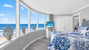 A bedroom with a curved window overlooking the sea, a bed with a blue and white patterned bedspread, and a television on the wall.