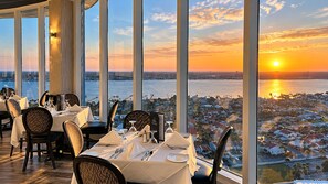 A dining area with tables set for a meal, large windows offering a view of a cityscape and sunset over water. - 2BR/2BA • Beachfront • Hot Tub • Pool • Ball Courts • Gym • Fire Pit • 8NE (Daytona Beach)