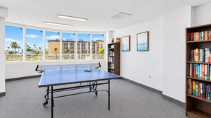 Ein Zimmer mit Tischtennisplatte, einer Bücherwand und großen Fenstern mit Blick auf Gebäude und Palmen.