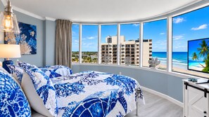 Une chambre d’hôtel dotée d’une baie vitrée incurvée, d’un lit avec une literie à motifs bleus et blancs, d’un bureau avec une télévision, et offrant une vue sur une plage et les bâtiments de l’hôtel.