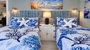 Ein Zimmer mit zwei Betten, jedes mit einem Meerstern-Kissen und einer blauen Bettdecke im Meeresstil. Ein Nachttisch mit Lampe und ein kleiner Tisch mit einem dekorativen Gegenstand sind vorhanden. Über dem Bett hängt ein Bild von einer Strandlandschaft.