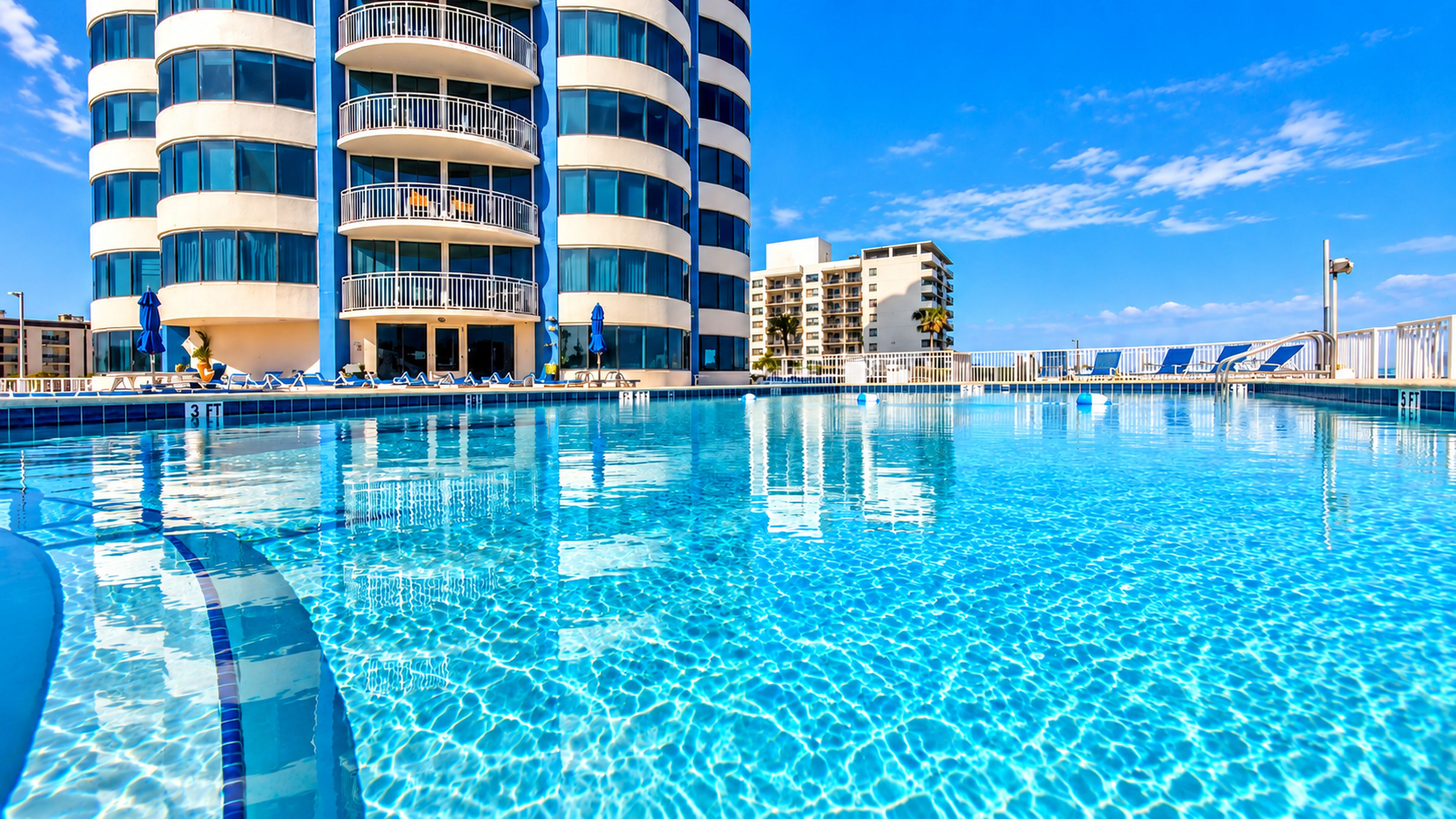 A swimming pool with clear blue water, surrounded by modern buildings with balconies and lounge chairs.