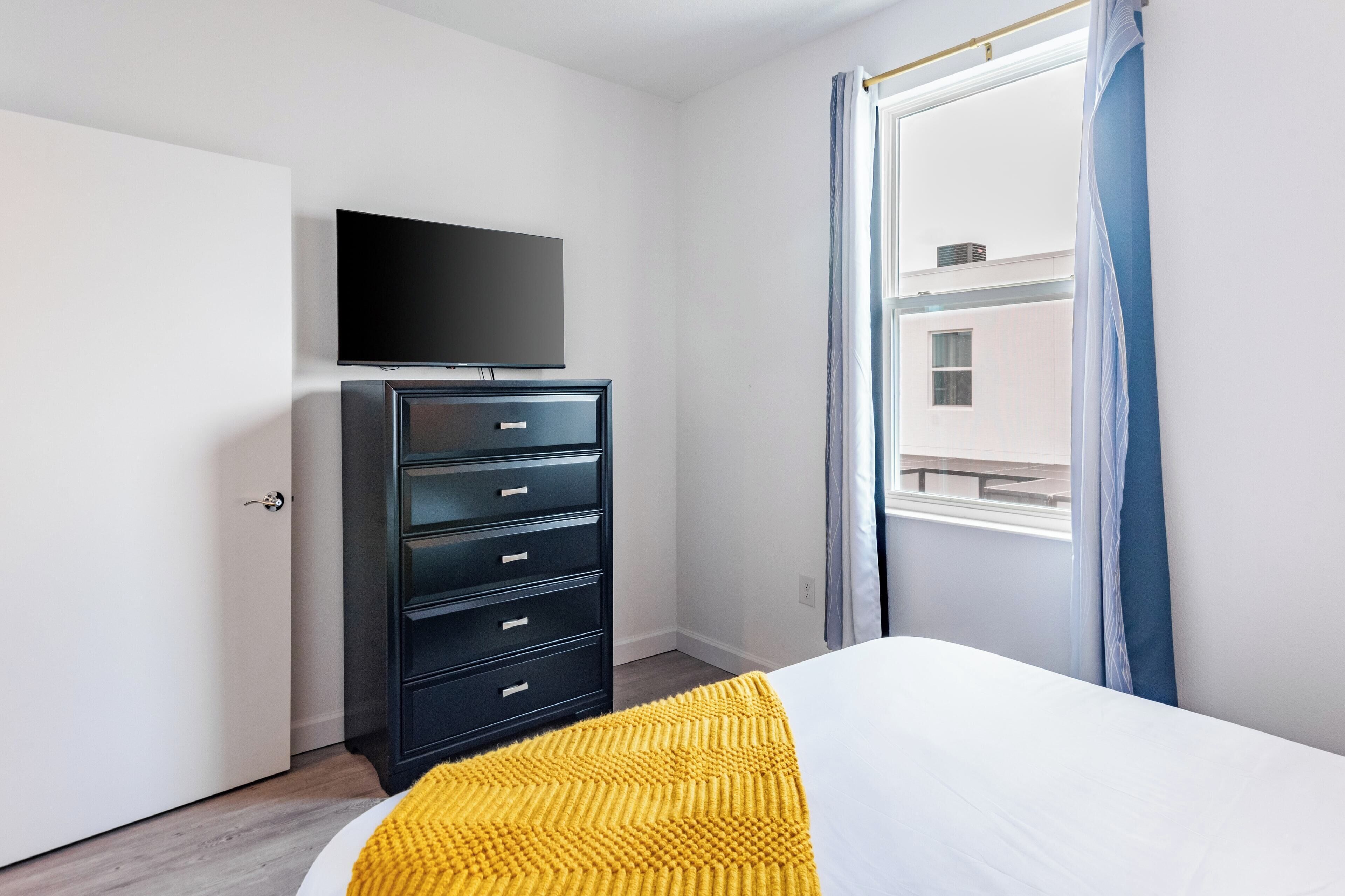 A hotel room with a bed, a flat-screen TV mounted on a dresser, a window with curtains, and a view of a building outside.
