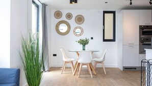 Una cocina moderna con armarios blancos, un comedor con una mesa redonda y sillas blancas, y un ventanal con cortinas.