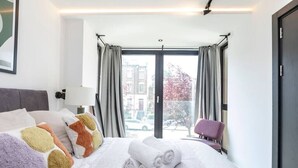 Habitación de hotel con cama, almohadas, una silla y vistas a edificios y árboles a través de amplias ventanas.