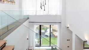 A modern interior with a glass staircase, a dining area with wooden tables, and a view of a garden with outdoor furniture.
