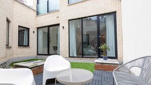 A modern outdoor patio with a brick wall, large sliding glass doors, and outdoor seating including white chairs and a round table.