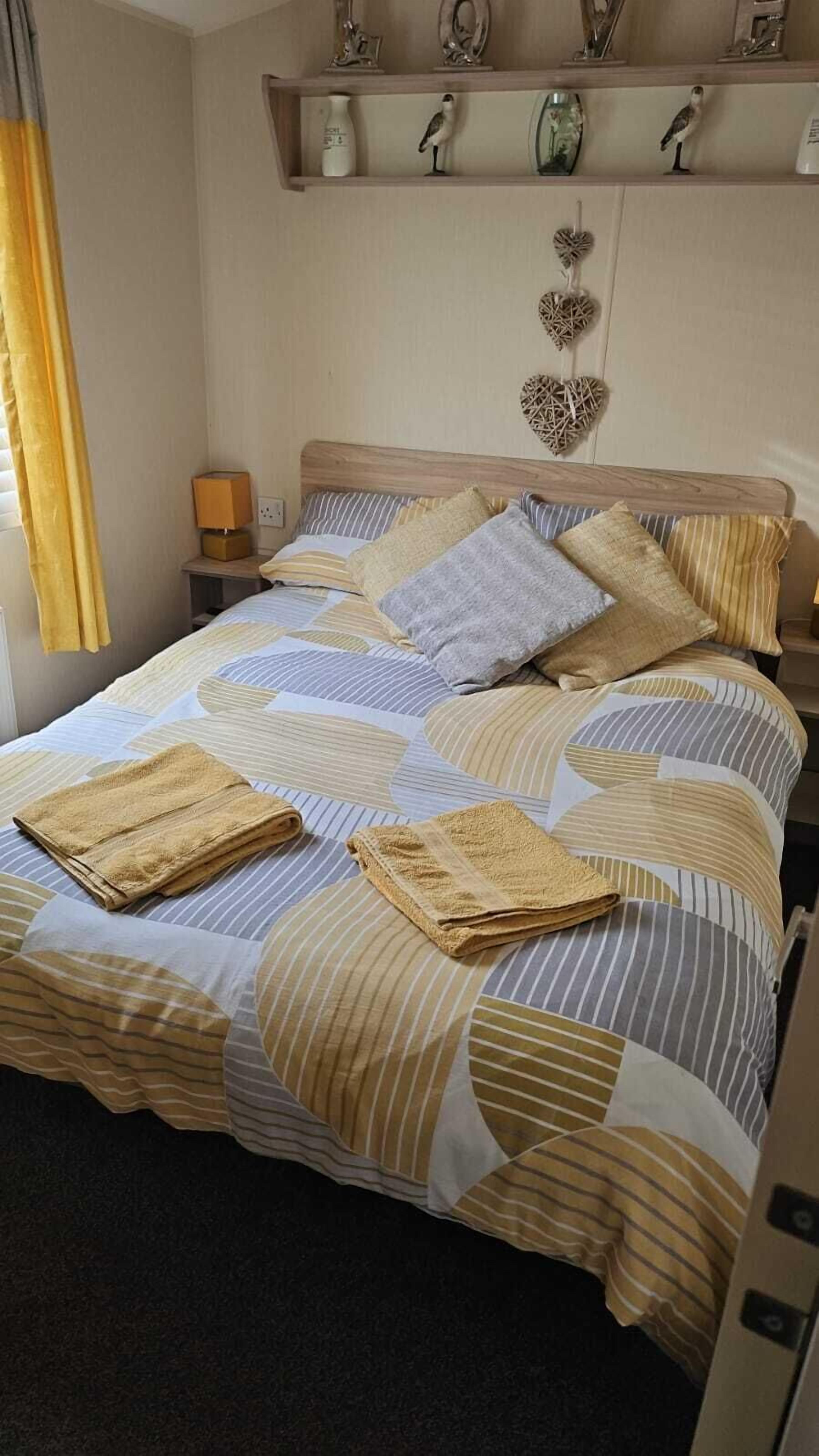 A neatly made bed with striped bedding and folded towels, a bedside lamp, and a shelf with decorative items.
