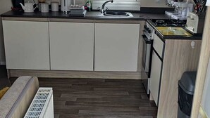 A compact kitchen with a sink, stove, and microwave. There is a window with blinds and a radiator on the floor. - 3 Bed Caravan - Sleeps 6 - Pets - Parking - Wifi (Skegness)