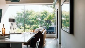 A modern interior with a dining area, a sofa, and large windows offering a view of trees and a building. - Kroger Lofts | Unit A | Rooftop Pool | Lake Views (Charlevoix)