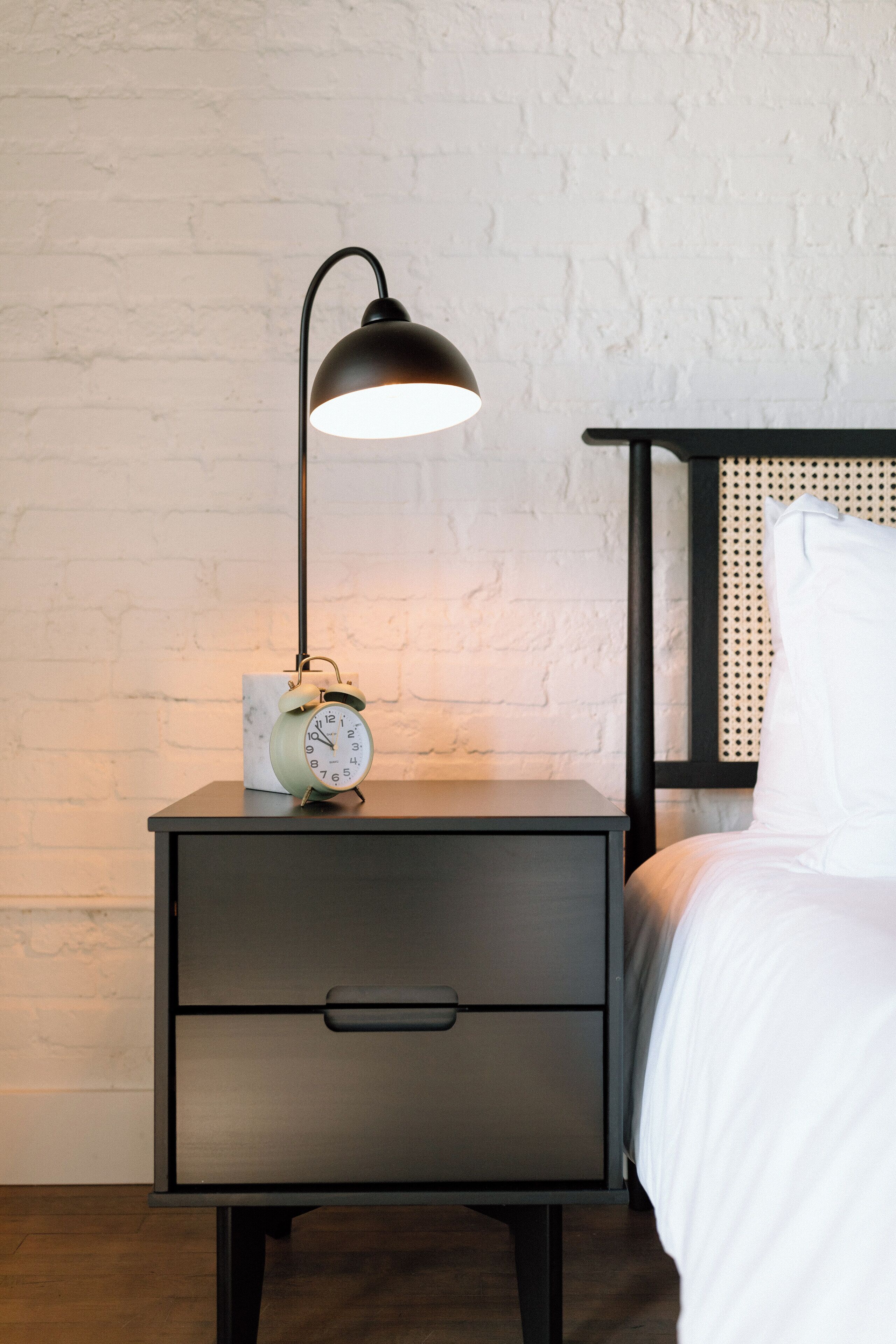 A nightstand with a lamp, an alarm clock, and a bed with a headboard.