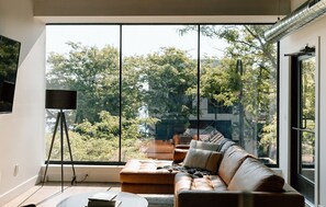 A modern living room with a large window, a sofa, a coffee table, and a floor lamp. - Kroger Lofts | Unit A | Rooftop Pool | Lake Views (Charlevoix)