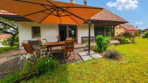 Une terrasse avec une table et des chaises sous un parasol, un petit jardin et une maison avec un toit en tuiles.