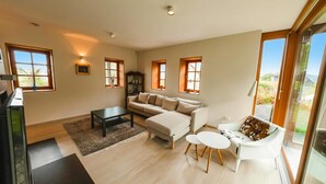 Un salon moderne avec un canapé, une table basse et une petite table à manger. De nombreuses fenêtres laissent entrer la lumière naturelle et offrent une vue sur l’extérieur.