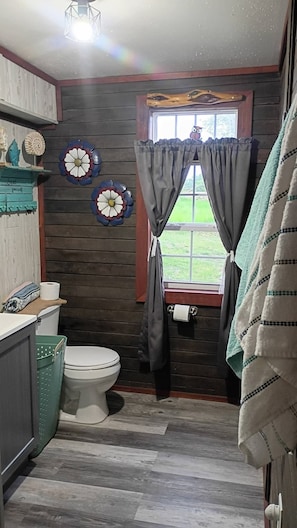 A bathroom with a toilet, a window with curtains, a wooden wall, and a towel rack.