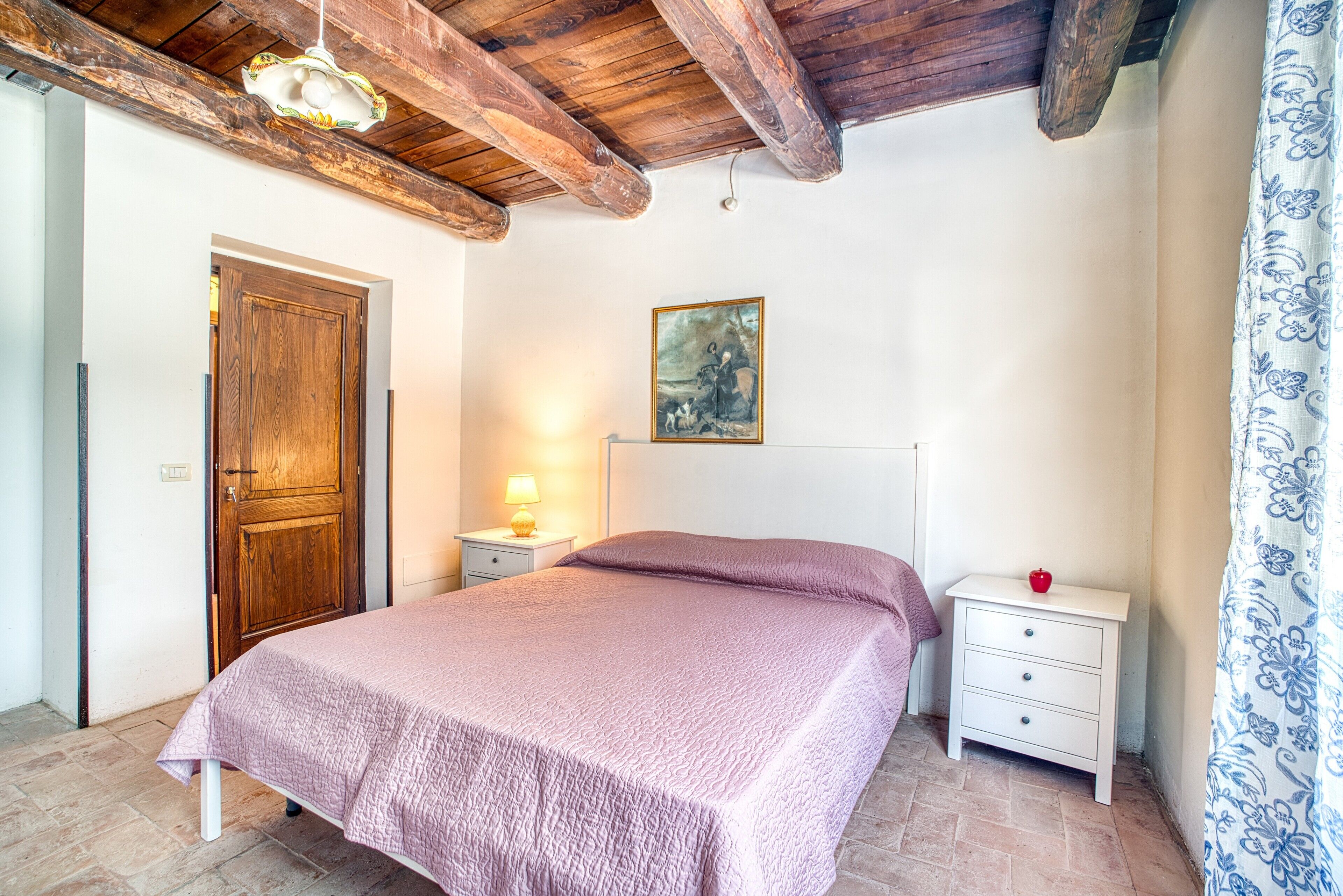 Ein Schlafzimmer mit Holzboden, einem Bett mit rosa Bettdecke, einem Nachttisch mit Lampe, einer weißen Kommode und einer Tür.