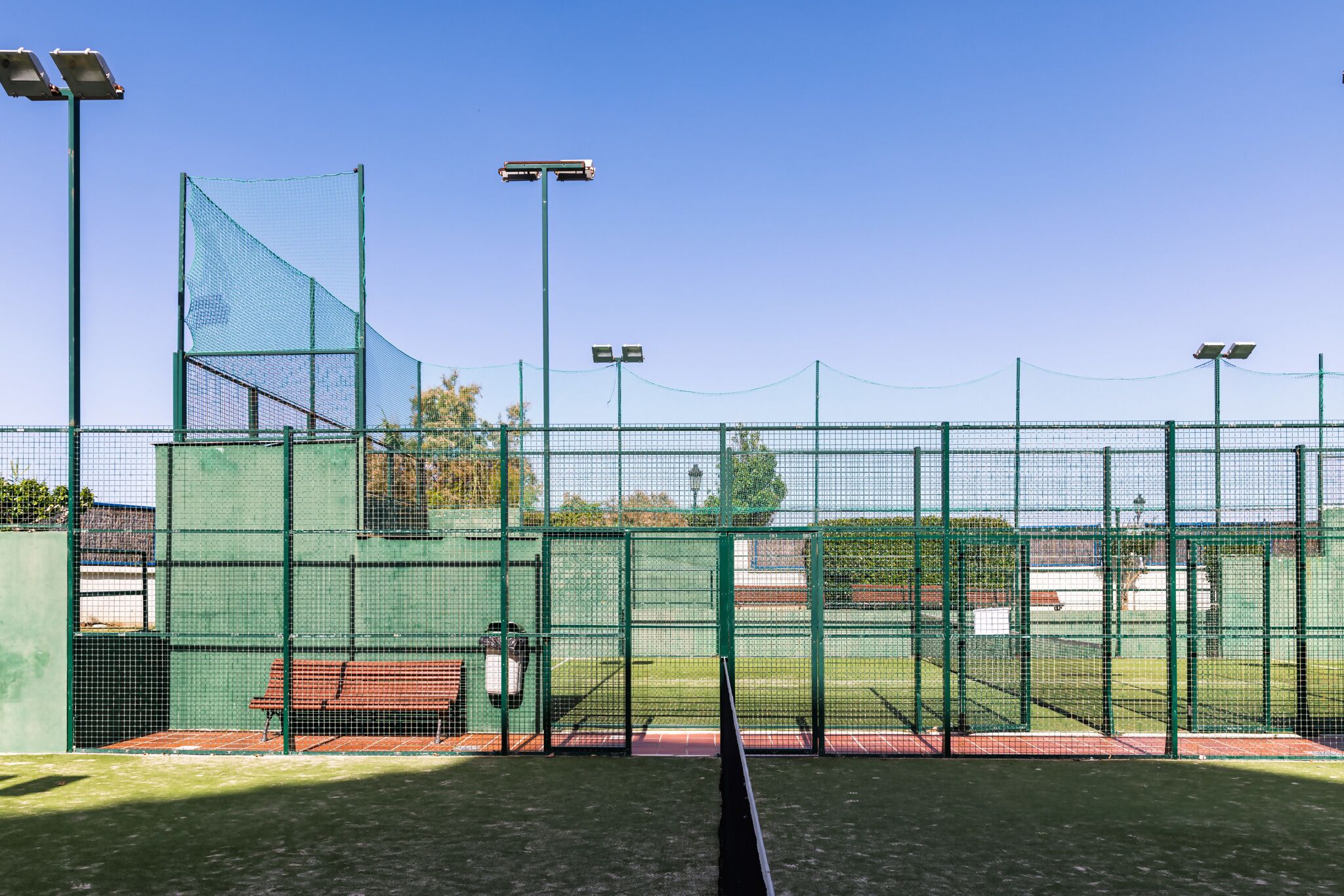 Tenniskenttä, jossa on vihreä aita, aurinkokennovalot ja penkki.