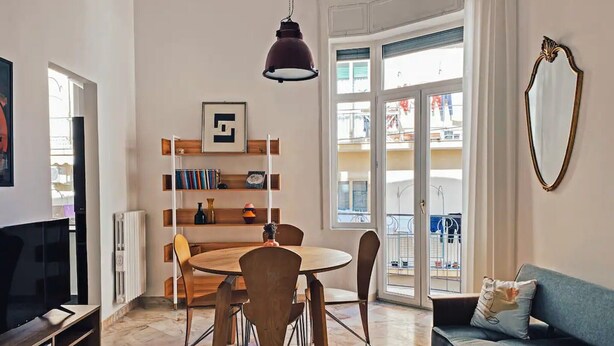 Een woonkamer met een televisie, een eettafel met stoelen, een boekenkast, een bank en een groot raam met uitzicht op het omliggende landschap.
