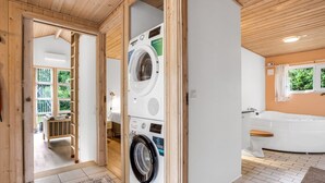 Een wasserette met een wasmachine en droger, een houten deur en een uitzicht op een slaapkamer met een bed en nachtkastje.