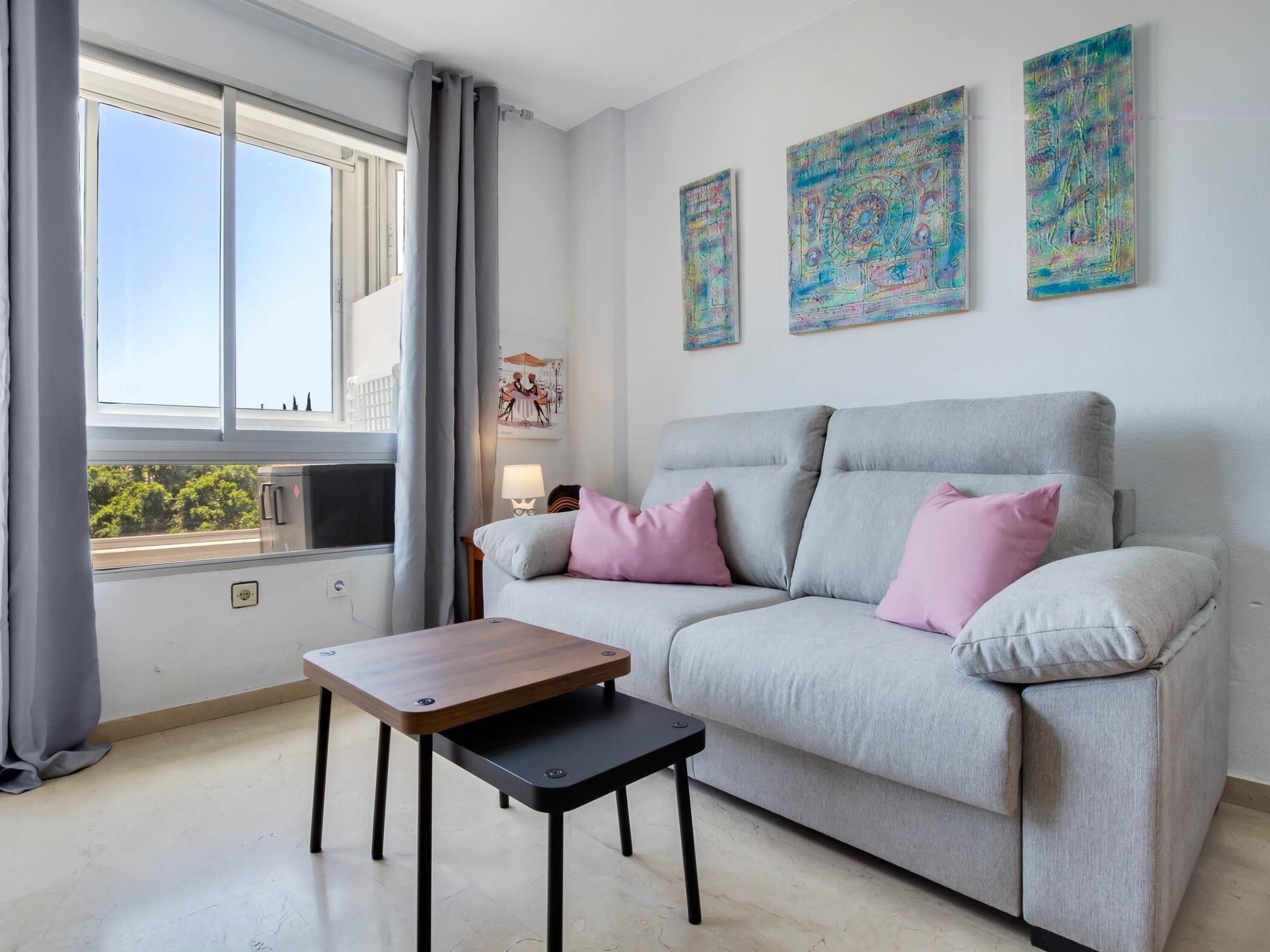 Uma sala de estar moderna com um sofá cinzento, dois almofadas cor-de-rosa, uma mesa de centro de madeira e pinturas abstratas nas paredes.